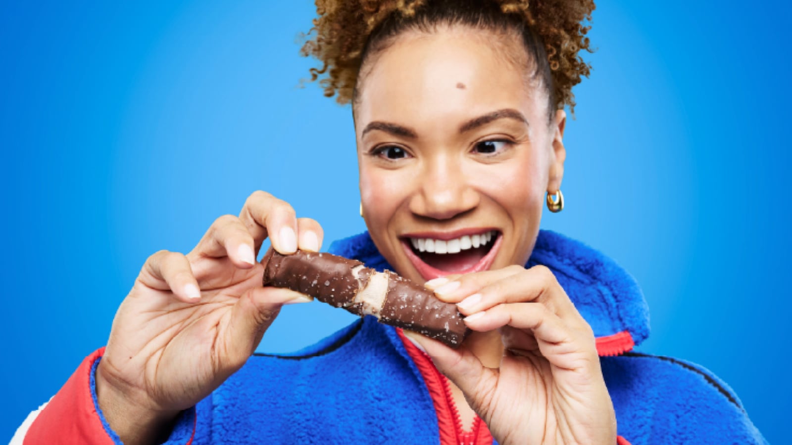 Smiling woman with curly hair pulled up, wearing a blue fleece jacket, pulling apart a chocolate-coated Built Puff protein bar against a bright blue background. The bar breaks to reveal a soft, marshmallowy interior.