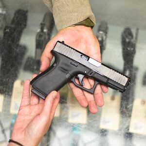 A salesman hands a woman a hand gun while he stands behind a showcase full of handguns in a shop in a gun shop.