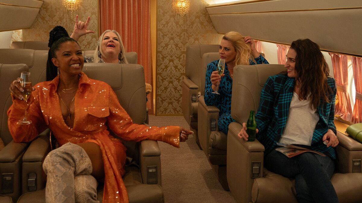 Renée Elise Goldsberry, Paula Pell, Busy Philipps, and Sara Bareilles raise glasses of champagne in a plane in a still from ‘Girls5Eva’