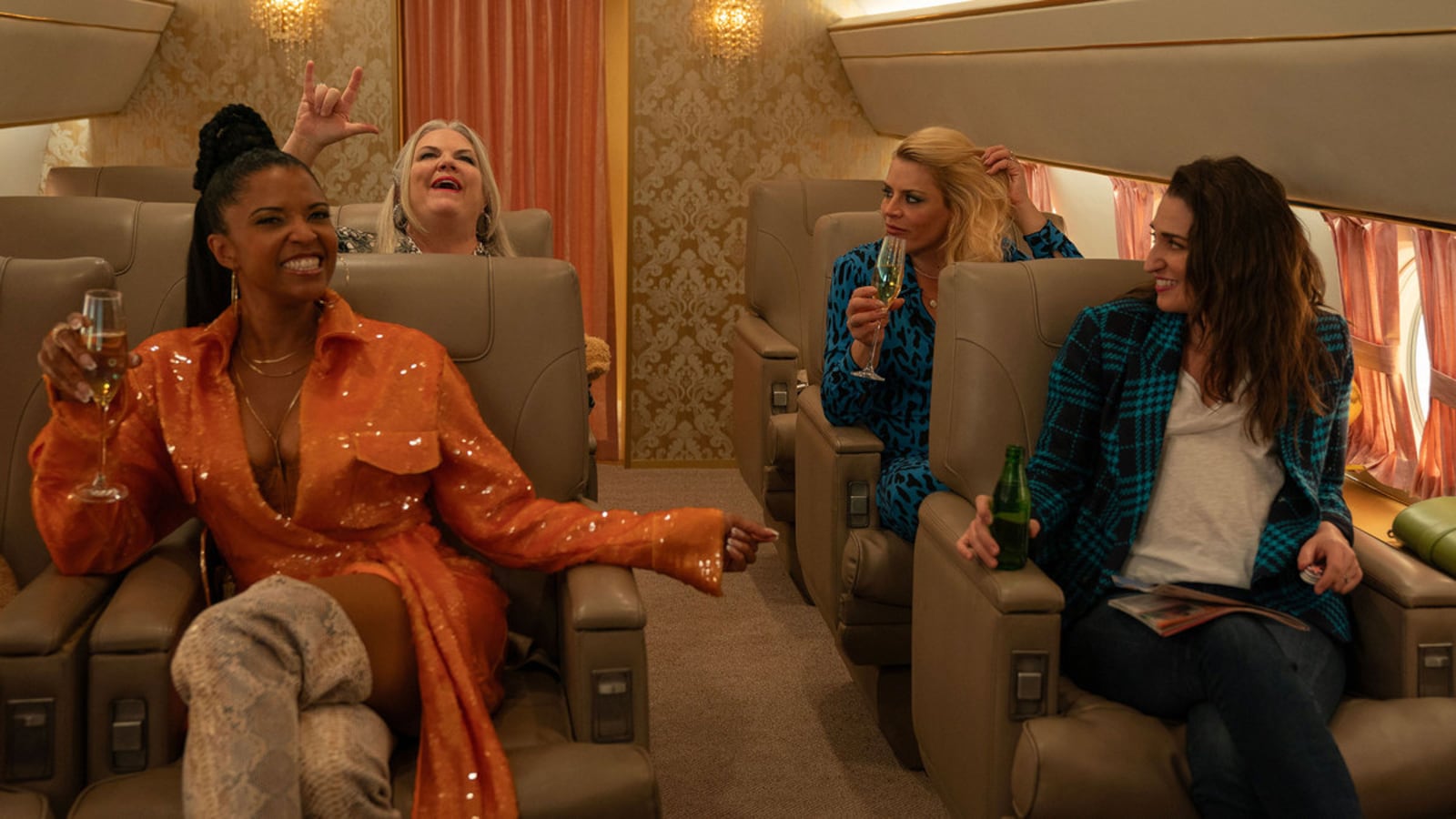 Renée Elise Goldsberry, Paula Pell, Busy Philipps, and Sara Bareilles raise glasses of champagne in a plane in a still from ‘Girls5Eva’