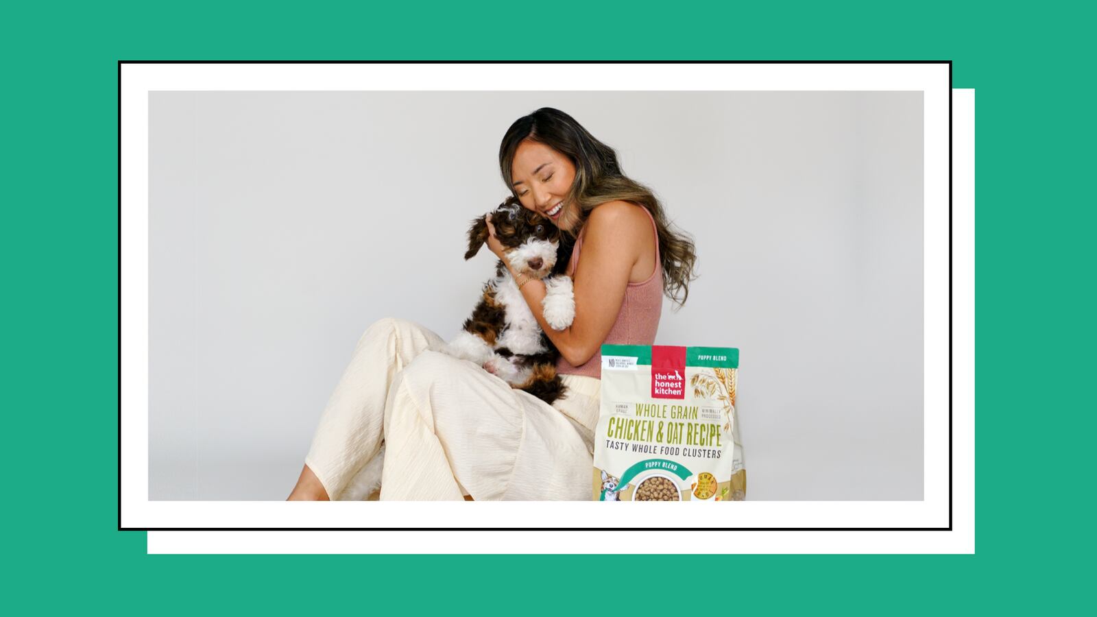 A woman with dark hair and wearing a pink shirt with white pants hugging a puppy. The puppy has white and black fur. In front of them is a bag of Grain Free Chicken Dry Dog Food.