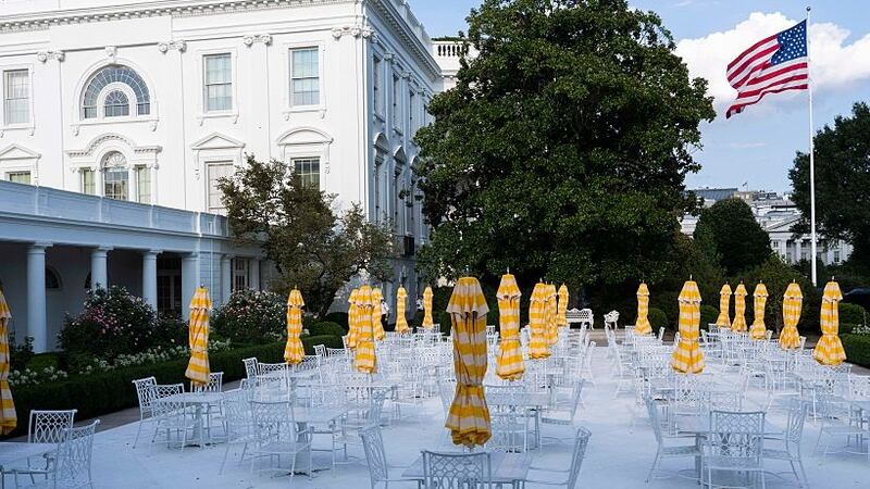 Metal tables and chairs along with yellow umbrellas decorate a paved over historic White House Rose Garden lawn.