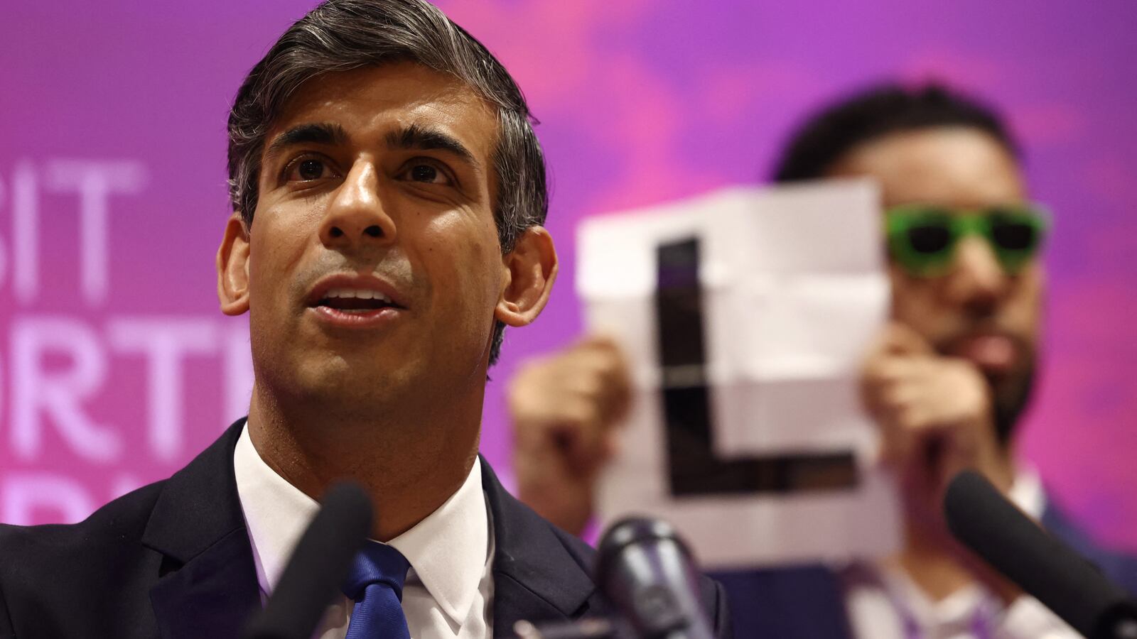 Independent candidate Niko Omilana holds an "L" behind Britain's Prime Minister and Conservative Party leader Rishi Sunak as he delivers a speech after retaining his seat as MP for Richmond and Northallerton in Northallerton, early on July 5, 2024.