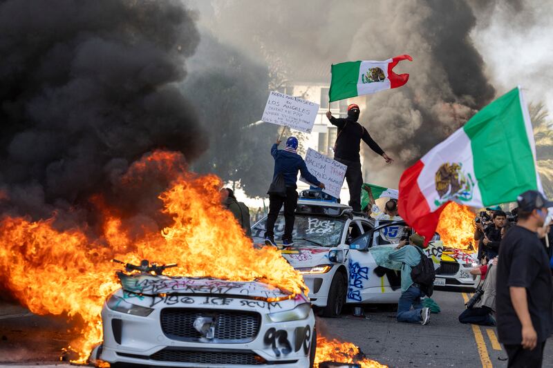 Demonstrators wave flags from atop a wrecked car, standing beside burning Waymo vehicles, as protesters clash with law enforcement in the streets surrounding the federal building during a protest following federal immigration operations in Los Angeles, California.