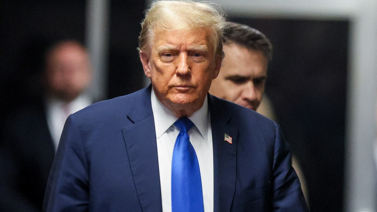 Donald Trump, wearing a suit, walks into court with a stoic look on his face.